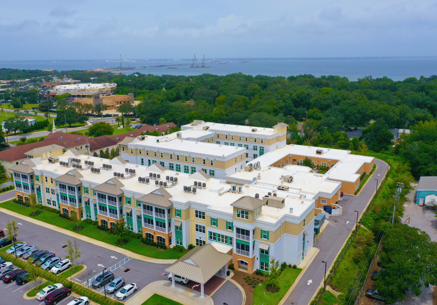 Aerial view of a multi-building apartment complex with a parking lot, edged by trees and a distant bay or ocean in the background.
