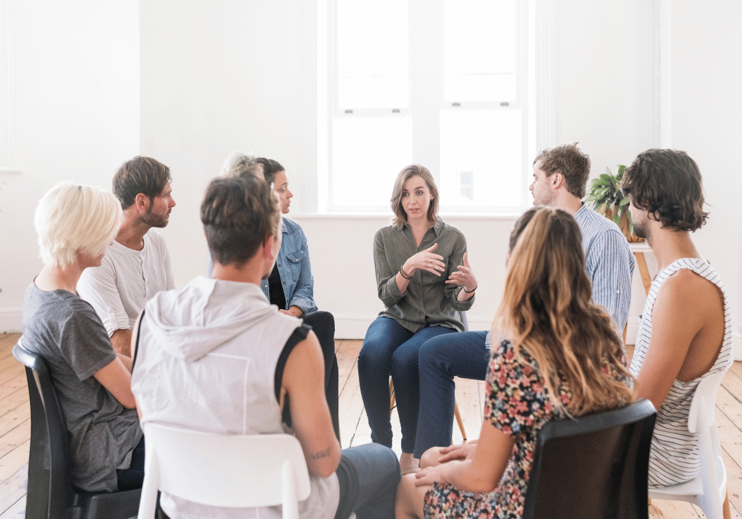 A woman speaks while seated in a circle of adults in a bright room, discussing together.