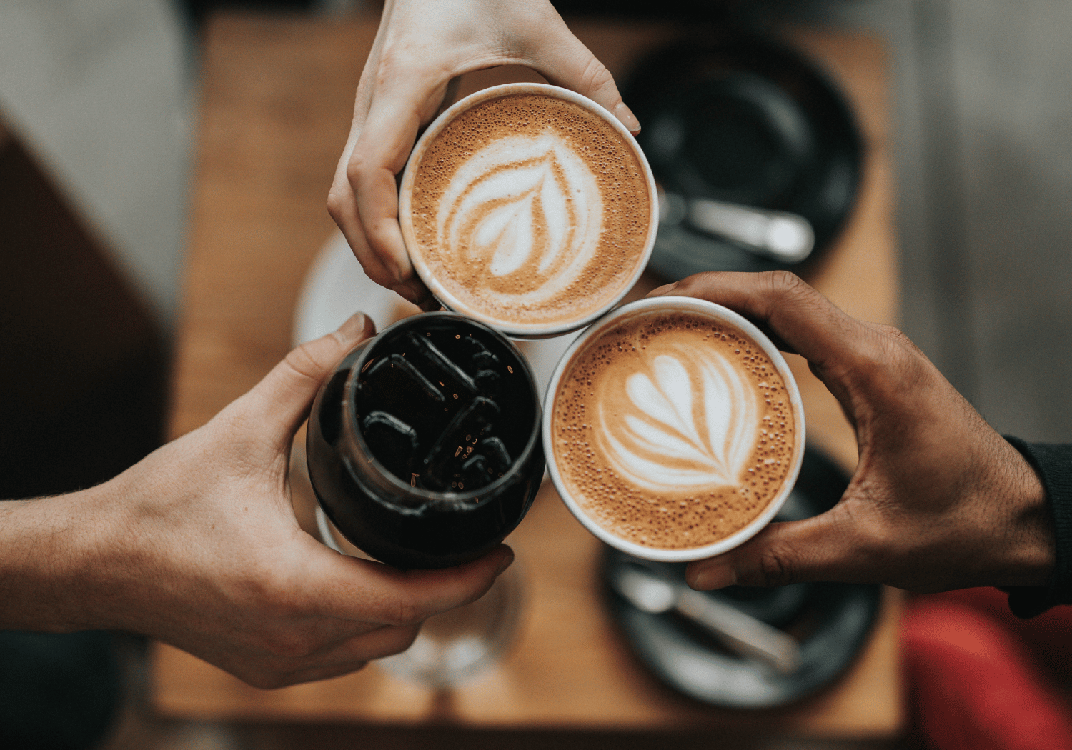 Three hands hold coffee cups in a circle over a wooden table, two with latte art leaves and one dark iced drink in a glass for a toast.